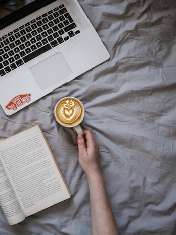 A hand holds a cup of latte art coffee on a gray bedspread. An open book lies nearby, alongside a laptop with a sticker on its surface. The scene conveys a cozy, relaxed atmosphere, suitable for reading or working.