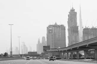 A bustling urban highway scene featuring several modern skyscrapers under construction, with cranes visible atop the tall buildings. The roadway has multiple lanes, with cars traveling in both directions. Overpasses and road signs are visible, and the scene is enveloped in a light haze, giving a slightly subdued appearance.