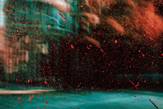 A glass surface covered with red paint splatters creates an abstract effect. The background is blurred, possibly a building or structure, seen through the paint-covered glass.