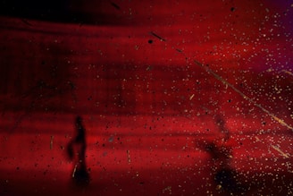 A dark, abstract scene with a deep red background and scattered, speckled textures throughout. Silhouetted figures are visible, adding a sense of mystery and depth. The background gradient shifts from red to hints of purple.