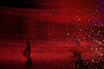 A dark, abstract scene with a deep red background and scattered, speckled textures throughout. Silhouetted figures are visible, adding a sense of mystery and depth. The background gradient shifts from red to hints of purple.