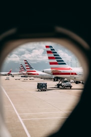 Multiple airplanes with distinctive red, white, and blue tail designs are parked on an airport tarmac, viewed through an oval window. The scene includes clear skies with a few clouds and numerous ground support vehicles such as baggage carts and trucks.