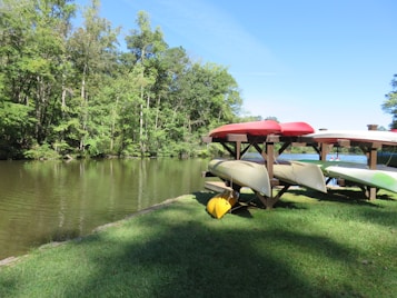 A serene lakeside setting with several canoes stacked neatly on wooden racks. The background features lush green trees and calm water reflecting the blue sky. The grass in the foreground is well-kept, adding to the tranquil atmosphere.