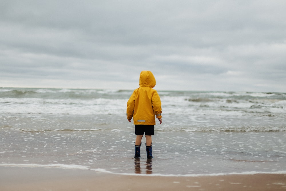 toddler wearing yellow hoodie and black shorts standing on seashore