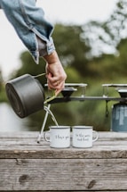 A hand in a denim jacket is pouring liquid from a black kettle into two white metal mugs placed on a wooden surface. The mugs have text on them; one says 'I love you' and the other reads 'to the moon and back'. In the background, a green, blurred natural setting can be seen.