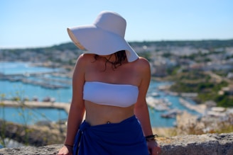 A person wearing a large white sun hat and a white bandeau top is standing in front of a scenic seaside view. The background features a coastal town with buildings and a harbor, with the sea and hills stretching into the distance. The person is also wearing a blue wrap or skirt.