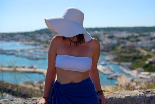 A person wearing a large white sun hat and a white bandeau top is standing in front of a scenic seaside view. The background features a coastal town with buildings and a harbor, with the sea and hills stretching into the distance. The person is also wearing a blue wrap or skirt.