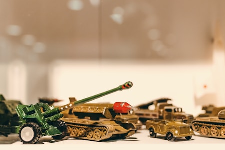 A collection of toy military vehicles, including tanks and trucks, is arranged in a display. The toys have a vintage look, with some painted in green and others in a muted gold color. The lighting creates a warm, nostalgic ambiance, highlighting the intricate details of the small models.