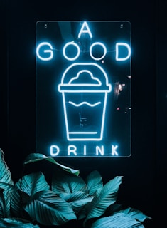 Neon sign featuring the words 'A GOOD DRINK' with an image of a drink cup. The sign emits a bright blue light against a dark background, with lush green leaves partially framing the bottom of the image.