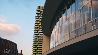An architectural scene featuring a modern glass building reflecting the sky and clouds, with an adjacent high-rise and industrial-looking structure in the background.
