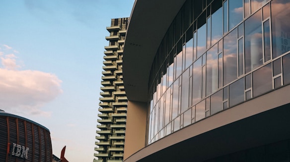An architectural scene featuring a modern glass building reflecting the sky and clouds, with an adjacent high-rise and industrial-looking structure in the background.