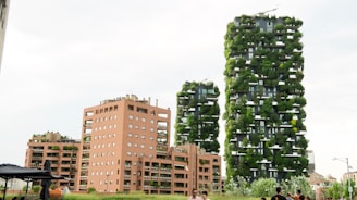 A modern urban skyline featuring two tall skyscrapers covered with abundant greenery, interspersed with terraces housing numerous plants. Adjacent to them are shorter buildings with terracotta facades and rooftop gardens. People are visible in the foreground, suggesting the scene is a populated urban environment.