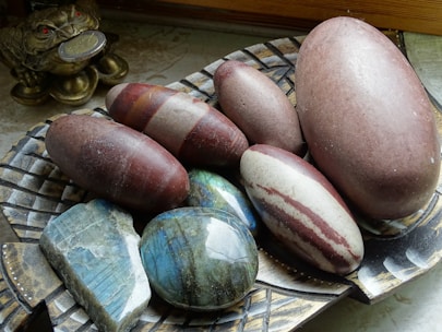 A collection of smooth, oval-shaped stones in various colors are displayed on a decorative, woven-patterned plate. There is a mixture of reddish-brown, grey, and striped stones with a glossy finish. To the side, a small, golden frog figurine holding a coin in its mouth is visible, adding an ornamental touch.