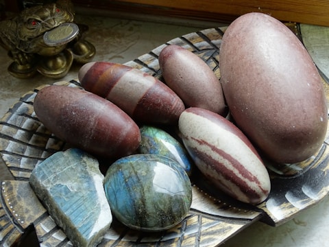 A collection of smooth, oval-shaped stones in various colors are displayed on a decorative, woven-patterned plate. There is a mixture of reddish-brown, grey, and striped stones with a glossy finish. To the side, a small, golden frog figurine holding a coin in its mouth is visible, adding an ornamental touch.