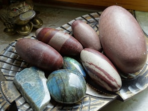 A collection of smooth, oval-shaped stones in various colors are displayed on a decorative, woven-patterned plate. There is a mixture of reddish-brown, grey, and striped stones with a glossy finish. To the side, a small, golden frog figurine holding a coin in its mouth is visible, adding an ornamental touch.