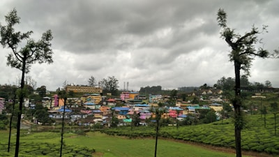 A small hillside town with colorful houses is nestled amid lush green vegetation. The area is surrounded by tea plantations and tall, sparse trees. Overhead, dark, cloudy skies suggest overcast weather.