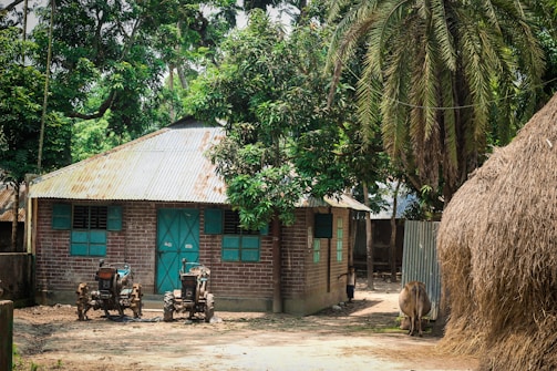A rural scene featuring a brick house with a corrugated metal roof and green window shutters, surrounded by lush green trees. Two small tractors or agricultural machinery are parked in front of the house. There is a large stack of hay or straw nearby, with a cow standing adjacent to it. The setting appears calm and tranquil.