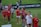 A group of children and pre-teens are walking on a grass field. Most are wearing red sports shirts, some with 'Washington Spirit' printed on them. They seem to be part of a sports event, possibly soccer, and look happy and excited. There is a banner in the background with a website URL.