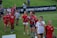 A group of children and pre-teens are walking on a grass field. Most are wearing red sports shirts, some with 'Washington Spirit' printed on them. They seem to be part of a sports event, possibly soccer, and look happy and excited. There is a banner in the background with a website URL.