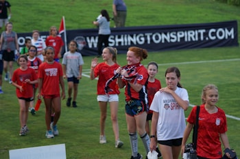 A group of children and pre-teens are walking on a grass field. Most are wearing red sports shirts, some with 'Washington Spirit' printed on them. They seem to be part of a sports event, possibly soccer, and look happy and excited. There is a banner in the background with a website URL.