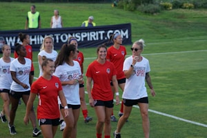 A group of women in sports attire are gathered on a grassy field, casually interacting and smiling. They are wearing shirts with 'Washington Spirit' written on them, suggesting they are part of a soccer team. A banner is visible in the background displaying the same name, reinforcing the team atmosphere. The image captures a moment of camaraderie and light-heartedness among the players.