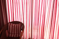 A wicker chair is positioned in front of a backdrop of red and white striped curtains. The chair contains a cushion that matches the pattern of the curtains, enhancing the visual harmony. Sunlight gently filters through the fabric, creating a warm and inviting atmosphere.