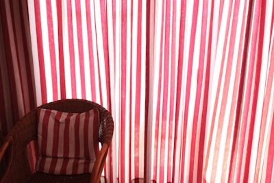 A wicker chair is positioned in front of a backdrop of red and white striped curtains. The chair contains a cushion that matches the pattern of the curtains, enhancing the visual harmony. Sunlight gently filters through the fabric, creating a warm and inviting atmosphere.