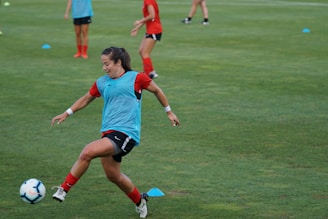 A soccer player wearing a red outfit and a blue training bib is in motion, about to kick a soccer ball on a grassy field. Other players can be seen in the background, also wearing red attire. The players appear to be practicing, with several training cones scattered across the field.