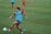 A soccer player wearing a red outfit and a blue training bib is in motion, about to kick a soccer ball on a grassy field. Other players can be seen in the background, also wearing red attire. The players appear to be practicing, with several training cones scattered across the field.