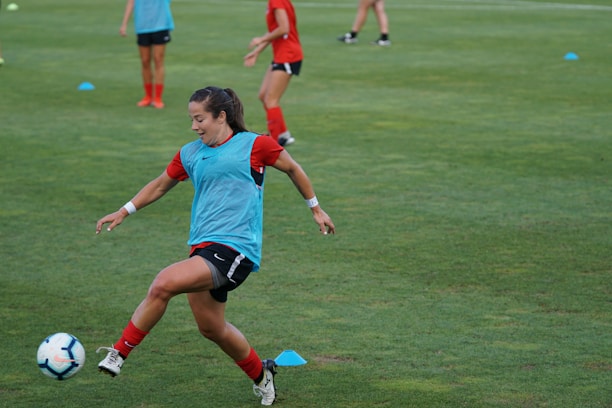 A soccer player wearing a red outfit and a blue training bib is in motion, about to kick a soccer ball on a grassy field. Other players can be seen in the background, also wearing red attire. The players appear to be practicing, with several training cones scattered across the field.