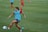 A soccer player wearing a red outfit and a blue training bib is in motion, about to kick a soccer ball on a grassy field. Other players can be seen in the background, also wearing red attire. The players appear to be practicing, with several training cones scattered across the field.