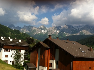 A picturesque alpine scene featuring traditional wooden houses with brown roofs, nestled in a lush green valley. In the background, a striking mountain range with rugged, snow-capped peaks rises dramatically into a partly cloudy sky.
