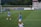 A soccer game scene on a green grass field with players wearing light blue jerseys and white shorts. A goalpost is visible in the background, and there are people sitting or standing on the bleachers. Advertising banners are displayed along the sidelines.