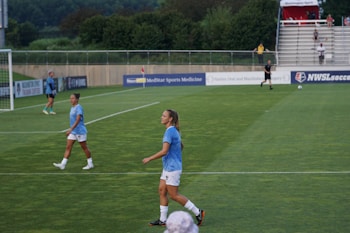 A soccer game scene on a green grass field with players wearing light blue jerseys and white shorts. A goalpost is visible in the background, and there are people sitting or standing on the bleachers. Advertising banners are displayed along the sidelines.