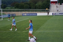 A soccer game scene on a green grass field with players wearing light blue jerseys and white shorts. A goalpost is visible in the background, and there are people sitting or standing on the bleachers. Advertising banners are displayed along the sidelines.