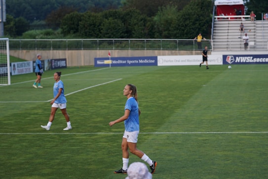 A soccer game scene on a green grass field with players wearing light blue jerseys and white shorts. A goalpost is visible in the background, and there are people sitting or standing on the bleachers. Advertising banners are displayed along the sidelines.