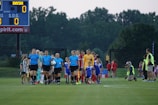 A group of referees and soccer players walks onto the field, accompanied by children serving as mascots. The referees are dressed in blue uniforms, and the players wear sports jerseys in red, yellow, and other colors. The scene takes place on a lush green grass field with a scoreboard in the background. The atmosphere suggests a professional or semiprofessional soccer match.