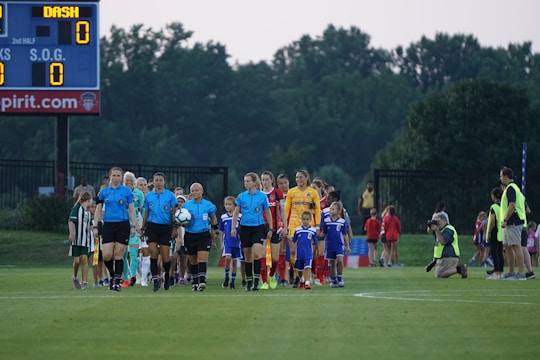 A group of referees and soccer players walks onto the field, accompanied by children serving as mascots. The referees are dressed in blue uniforms, and the players wear sports jerseys in red, yellow, and other colors. The scene takes place on a lush green grass field with a scoreboard in the background. The atmosphere suggests a professional or semiprofessional soccer match.