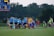 A group of referees and soccer players walks onto the field, accompanied by children serving as mascots. The referees are dressed in blue uniforms, and the players wear sports jerseys in red, yellow, and other colors. The scene takes place on a lush green grass field with a scoreboard in the background. The atmosphere suggests a professional or semiprofessional soccer match.