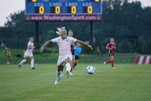 A soccer player in a white uniform is kicking a ball on a grassy field. Behind her, other players are in action, wearing different team colors. In the background, there is a scoreboard displaying statistics. The game appears to be taking place outdoors, with a fenced area and trees in the distance.