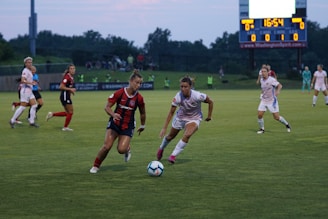 Two soccer players intensely compete for the ball on a grassy field, one wearing a red and black jersey and the other in a white uniform. The background shows more players, a scoreboard displaying a tied game, and a sparsely populated spectator area.