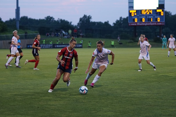 Two soccer players intensely compete for the ball on a grassy field, one wearing a red and black jersey and the other in a white uniform. The background shows more players, a scoreboard displaying a tied game, and a sparsely populated spectator area.