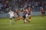 A women's soccer game in progress with players actively contesting for the ball. A player in a white jersey and another in a red and blue jersey are about to make contact with the ball, surrounded by teammates and opponents. The background shows a large crowd of spectators in a stadium setting.