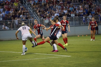 A women's soccer game in progress with players actively contesting for the ball. A player in a white jersey and another in a red and blue jersey are about to make contact with the ball, surrounded by teammates and opponents. The background shows a large crowd of spectators in a stadium setting.