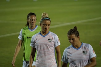 Three female athletes wearing sports jerseys are walking on a grassy field. The central figure is wearing a striped white and blue shirt with the 'DASH' logo, and her hair is tied up in a bun. To her right, another athlete has a similar shirt, with short hair tied back. The third person is wearing a green vest over her white shirt. The background is slightly blurred, focusing on the athletes in the foreground.