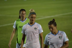Three female athletes wearing sports jerseys are walking on a grassy field. The central figure is wearing a striped white and blue shirt with the 'DASH' logo, and her hair is tied up in a bun. To her right, another athlete has a similar shirt, with short hair tied back. The third person is wearing a green vest over her white shirt. The background is slightly blurred, focusing on the athletes in the foreground.
