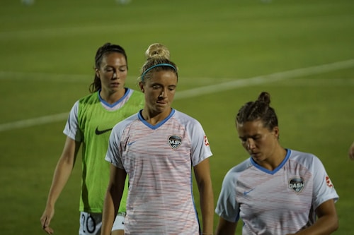 Three female athletes wearing sports jerseys are walking on a grassy field. The central figure is wearing a striped white and blue shirt with the 'DASH' logo, and her hair is tied up in a bun. To her right, another athlete has a similar shirt, with short hair tied back. The third person is wearing a green vest over her white shirt. The background is slightly blurred, focusing on the athletes in the foreground.