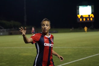 A soccer player wearing a red and blue jersey with 'GEICO' on it is on the field during a night game, gesturing with her hand. The background shows a scoreboard illuminated in the distance and another player in a yellow kit.