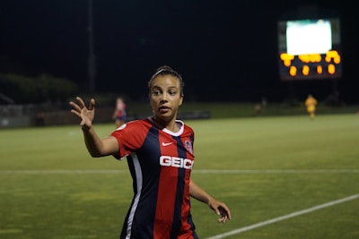 A soccer player wearing a red and blue jersey with 'GEICO' on it is on the field during a night game, gesturing with her hand. The background shows a scoreboard illuminated in the distance and another player in a yellow kit.