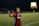 A soccer player wearing a red and blue jersey with 'GEICO' on it is on the field during a night game, gesturing with her hand. The background shows a scoreboard illuminated in the distance and another player in a yellow kit.